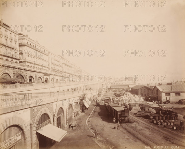 Current Boulevard Ernesto Che Guevara, Algiers, Algeria, photograph, View of the boulevard and the freight transshipment below., inscription: '1748. ALGER LE QUAI.', c. 1866 - c. 1890, photographer, paper, pasteboard, albumen print, height 213 mm, width 265 mm x width 440 mm, height 324 mm, North Africa, A wide riverside scene showing a long row of multistory buildings with arched lower levels and balconies along a raised promenade, a roadway and rail tracks running beside them, freight cars on the tracks, stacks of large sacks and barrels piled near warehouse-like sheds with pitched roofs, scattered carts and crates, several small groups of people and blurred figures moving along the road, lamp posts and railings along the elevated walkway, and distant low buildings and arches fading into the background.
