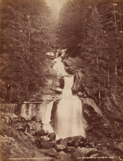 Waterfall, Triberg, Germany, photograph, Triberg as stated in the inscription should officially be: Triberg im Schwarzwald. It is a place in the German state of Baden-Württemberg and is best known for the Triberg Waterfalls. These waterfalls (7 in total, combined length 163 meters) are among the highest in Germany., inscription: 'JK BERBERICH TRIBERG 1892', inscription: 'TRIBERG DER WASSERFALL TOTAL', photographer: JK Berberich, 1892, Baden-Württemberg, pasteboard, paper, albumen print, albumendruk, height 274 mm, width 212 mm x width 329 mm, height 433 mm, A vertical sepia photograph showing a multi-tiered waterfall flowing down a rocky ravine surrounded by tall evergreen trees, with water cascading over large boulders and forming smooth white streams; a simple wooden bridge spans the ravine near the upper section of the falls, and scattered rocks and tree trunks line the foreground and sides of the scene.