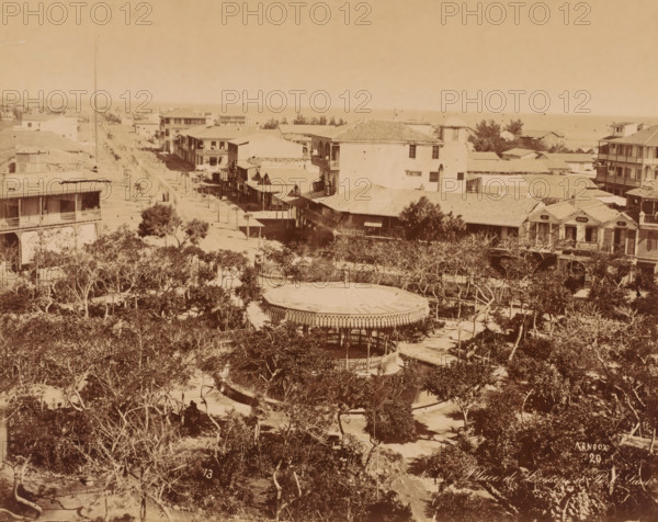 Place de Lesseps, Port Said, Egypt, photograph, Egypt., signature: 'Arnoux', inscription: 'No ? Place de Lesseps a Port Said', signature: 'Arnoux, 20', photographer: Hippolyte Arnoux, c. 1860 - c. 1890, pasteboard, paper, albumen print, height 213 mm, width 269 mm x width 467 mm, height 324 mm, North Africa, A wide sepia-toned view of a coastal town with rows of two-story buildings and covered porches lining a sandy street that leads toward the distant shoreline, a bandstand with a decorative roof and railing sits in a tree-filled park in the foreground surrounded by pathways and scattered trees, various tiled and pitched roofs rise behind the park, balconies and verandas are visible on several buildings, a few small canopies extend over sidewalks, trees cast dappled shadows across the scene, and the horizon shows a strip of sea meeting the sky.