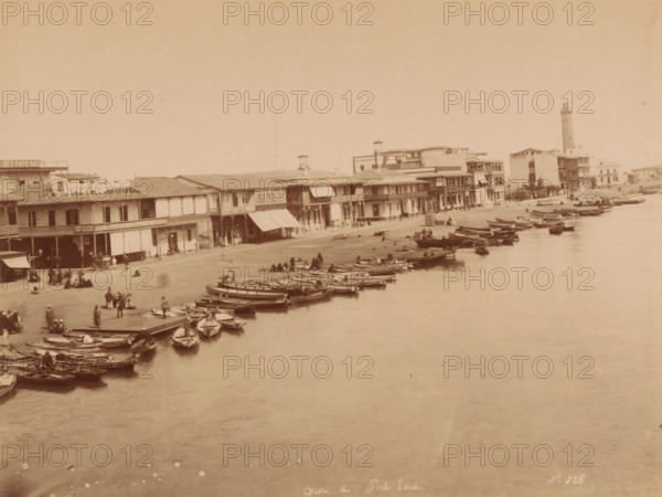 Quai de Port Said, Egypt, photograph, Egypt., inscription: 'quai de Port Said No. 326', photographer: attributed to Hippolyte Arnoux, c. 1860 - c. 1891, Attributed to, pasteboard, paper, albumen print, height 210 mm, width 278 mm x width 459 mm, height 323 mm, North Africa, Port Said, A waterfront scene with a row of two-story buildings along a shoreline, awnings and covered balconies on some facades, a lighthouse tower visible toward the right, numerous small wooden boats moored along the water's edge and floating on calm water, groups of people standing and sitting on the shore and near the boats, a wide expanse of water in the foreground and an open sky above.