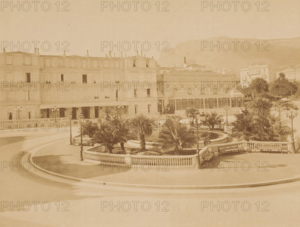 Square with roundabout with palm trees and pond in the middle and houses around it, photograph, Square with roundabout with palm trees and pond in the middle and houses around it., inscription: '622 ...', inscription: 'Phot. ND', signature: 'ND Phot.', photographer: Étienne Neurdein, c. 1880 - c. 1900, pasteboard, paper, albumen print, height 210 mm, width 276 mm x width 474 mm, height 323 mm, A wide view of an ornate building facade with multiple rows of windows and balconies adjacent to a structure with large arched windows, set behind a curved stone balustrade enclosing a landscaped area with palm trees, trimmed shrubs, and a circular water feature. Decorative street lamps with multiple globes line the paved terraces and paths, and benches sit along the balustrade. In the foreground a broad paved promenade sweeps in curved bands, and in the background low hills rise behind the buildings. The image has a uniform sepia tone.