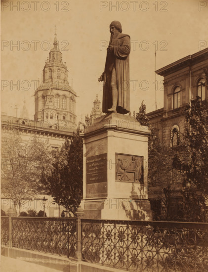 Gutenbergdenkmal and the Cathedral, Mainz, Germany, photograph, Germany., photographer: K, c. 1880 - c. 1900, pasteboard, paper, albumen print, height 269 mm, width 207 mm x width 357 mm, height 419 mm, Rhineland-Palatinate, A tall stone statue of a robed figure stands on a rectangular pedestal, the figure holding a small object in one hand and wearing a long cloak and head covering, with a long beard visible; the pedestal features a carved relief panel showing two seated figures at a table and another inset plaque; ornate metal railings run along the foreground in front of the pedestal; leafy trees partially obscure the base of the monument and a multi-story stone building with arched windows is visible to the right; in the background a large ornate tower with layered architectural details rises against the sky.