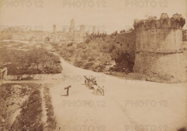 Panorama of San Gimignano, Tuscany, photograph, Cityscape with a road and an old fortress, city wall in the foreground on the right, and on the road people with horse and cart and stacks of wood on their shoulders. San Gimignano is known for its thirteen towers that dominate the city's silhouette, some of which can be seen here. These were erected in the 12th and 13th centuries by rival wealthy families. The towers served as residences and fortifications, but above all as objects of prestige. At its peak, the city had about 70 towers., inscription: 'No. 6378 San Gimignano - Panorama', inscription: 'NORWAY', c. 1870 - c. 1900, photographer, pasteboard, paper, albumen print, height 190 mm x width 250 mm, height 444 mm x width 356 mm, San Gimignano, A wide sepia-toned view of a town and surrounding landscape dominated on the right by a tall stone fortification with a rounded bastion topped by vegetation, a long curving stone wall extending inward and bordered by trees and scrub, and on th...