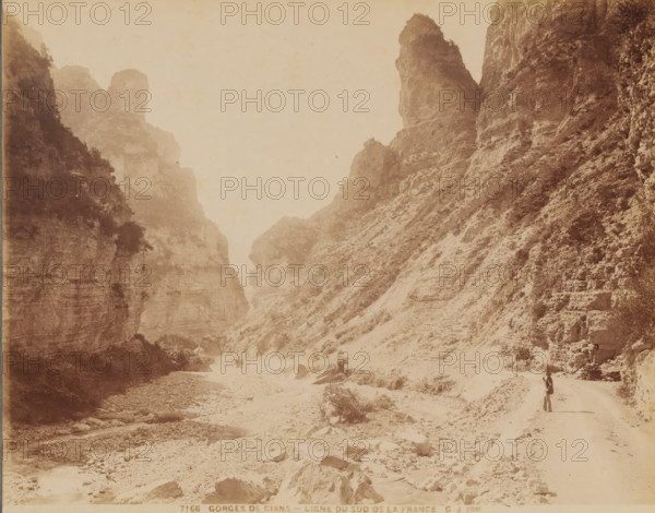 Ligne du Sud de la France, Gorges de Cians', France, photograph, inscription: '7166. GORGES DE CIANS - LIGNE DU SUD DE LA FRANCE', inscription: 'GJ photo.', photographer: Jean Gilletta, c. 1880 - c. 1900, Jean Gilletta, photographer, paper, pasteboard, albumen print, height 217 mm, width 275 mm x width 317 mm, height 387 mm, A sepia-toned scene of a narrow rocky canyon with steep cliff faces on both sides, showing layered rock textures and sparse vegetation clinging to the slopes, a dry rocky riverbed running through the canyon floor with scattered boulders and small pools of water, a winding dirt path along one side of the canyon, and a solitary person standing on the path carrying an object over their shoulder.