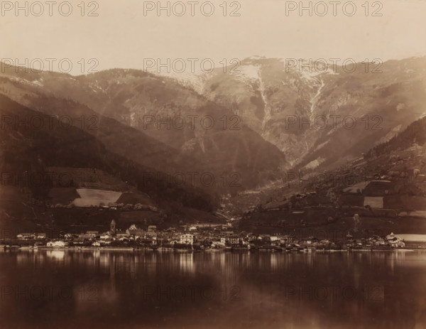 Landscape with a lake and mountains and a place on the shore of the lake, photograph, c. 1880 - c. 1900, photographer, pasteboard, paper, albumen print, height 205 mm, width 265 mm x width 373 mm, height 300 mm, A lakeside town with clustered buildings along the shore reflected in calm water, set against steep, forested hills and high mountains with patches of snow and visible gullies, terraces and scattered farm plots on the slopes, and a sky above.