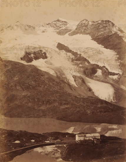 Bernina Pass, Switzerland, photograph, The Bernina Pass connects the Veltlin with the Engadin., inscription: 'Cauma See (Flims)', number: '21', c. 1890 - c. 1910, photographer, pasteboard, paper, albumen print, albumendruk, height 266 mm, width 205 mm x width 300 mm, height 379 mm, A wide mountain scene shows a steep rocky foreground leading up to snow-covered ridges and glaciers with visible crevasses and dark exposed rock bands, a pale sky above, a reflective body of water in the lower middle reflecting parts of the landscape, a curved narrow bridge crossing the water, and a multi-story pale building with a pitched roof and several small outbuildings situated near the water's edge.