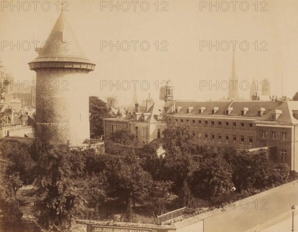 Cityscape with (water) tower and cathedral, France ?, photograph, France ?., c. 1880 - c. 1900, photographer, pasteboard, paper, albumen print, height 215 mm, width 275 mm x width 400 mm, height 318 mm, A wide sepia-toned cityscape showing a large round stone tower with a conical roof on the left, rows of multi-story buildings with pitched roofs and dormer windows to the right, a smaller square tower with a domed top and louvered openings near the center, distant slender spires on the horizon, a foreground area filled with dense trees and shrubs, a low wall and railing along the bottom edge, and a solitary streetlamp near the lower right corner.