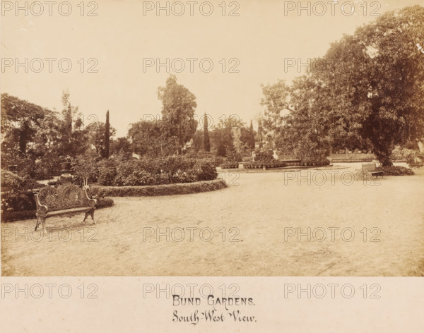 Bund Gardens, Poona, India, photograph, View of a park in Poona, also known as Pune en Puna and formerly called Punavadi. This is an Indian city not far from Bombay in the Western Ghats. The city is located in the district of the same name, Poona., inscription: 'Bund Gardens, South-West View', inscription: 'BUND GARDENS, POONA., South-West View.', c. 1875 - c. 1900, photographer, paper, pasteboard, albumen print, height 155 mm, width 238 mm x width 357 mm, height 278 mm, A wide sepia photograph of a garden scene with a gravel foreground leading to planted beds edged with low hedges and rows of potted plants, ornate cast-iron benches with decorative backs placed near the paths, large leafy trees including a very large tree on the right, taller slender trees and varied shrubs filling the midground, a circular raised planting area with a statue and surrounding pots, and a distant backdrop of more trees under an open pale sky.