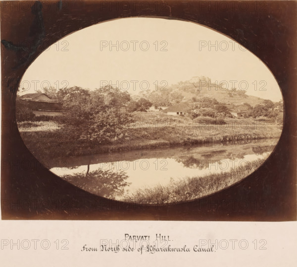 Parvati Hill from the north side of Rharakwasla Canal, India, photograph, India. The photo is elongated oval., inscription: 'Bund Gardens, South-West View', inscription: 'BUND GARDENS, POONA., South-West View.', c. 1875 - c. 1900, photographer, paper, pasteboard, albumen print, albumendruk, height 182 mm x width 246 mm, height 304 mm x width 393 mm, An oval-framed sepia landscape showing a hill with a cluster of trees and small buildings on its slopes, a body of water in the foreground reflecting the vegetation and hillside, grassy banks along the water, scattered trees and shrubs across the midground, and a dark vignette around the oval frame.