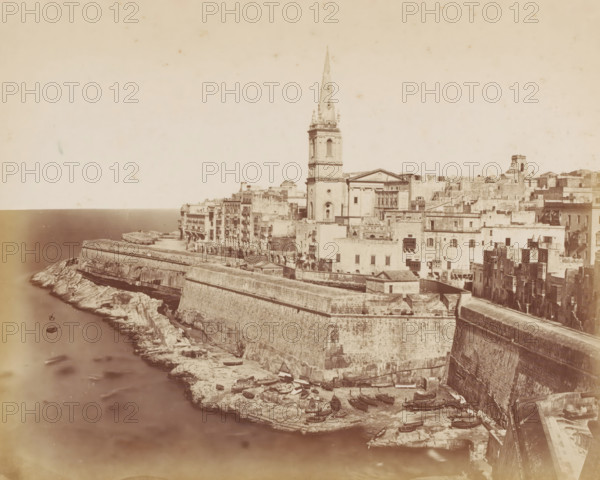 Malta, photograph, Malta is a dwarf state located in the Mediterranean Sea consisting of the islands of Malta, Gozo, Comino and the small uninhabited islands of Manoel Island, Cominotto, Filfla, Fungus Rock and the Saint Paul's Islands., inscription: 'Photo: Micallef e Borg ?', inscription: 'Malta', c. 1880 - c. 1900, photographer, paper, pasteboard, albumen print, albumendruk, height 199 mm, width 250 mm x width 359 mm, height 264 mm, A coastal cityscape seen from above showing a stone seawall and rocky shoreline with small boats pulled up on the rocks, a densely packed cluster of light-colored stone buildings rising behind the wall, and a prominent church with a tall slender spire standing among the buildings, all under a pale sky.