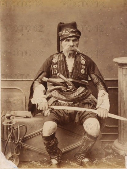 Portrait of old man in traditional costume, dervish?, photograph, dervish?., c. 1880 - c. 1900, photographer, paper, pasteboard, albumen print, height 228 mm, width 173 mm x width 389 mm, height 295 mm, An older man is seated facing forward, wearing a decorated dark jacket with patterned trim and a striped cloth wrapped around his waist, a tall hat with a long tassel, and high embroidered boots; he has a large bushy mustache and holds a curved sword horizontally across his lap with both hands, while several ornamental daggers or short blades are tucked into his waist sash and a coiled hose-like object and metal fixture sit beside the bench he occupies.