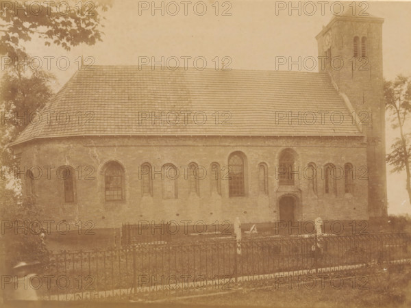 View of the Johanneskerk in Weidum, photograph, annotation: 'Weidum, Weidum, Baaderadeel', annotation: 'NH Church in Weidum (Friesland), list 1902, I. 278 No. 1394, Baarderadeel', photographer: AJM Mulder (possibly), 1902, Attributed to, paper, pasteboard, albumen print, albumendruk, height 168 mm x width 225 mm, church (exterior), Johanneskerk (Weidum), A large brick building with a steep tiled roof and a square tower is visible, showing a row of arched windows and a single wooden door set into the wall, with masonry details and weathering on the brick surface; a wrought iron fence runs across the foreground enclosing a grassy area with a few upright stone markers and a small fenced plot, while trees and overhanging branches frame the scene at the top and sides and a blurred figure appears near the lower corner of the image.