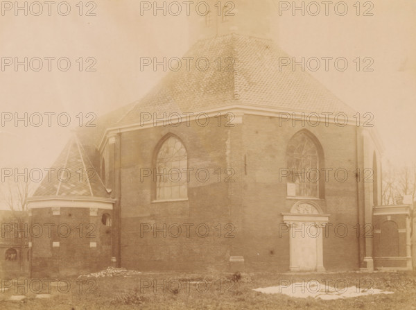 West side of the Van Harenskerk in Sint-Annaparochie, photograph, annotation: 'NH Church in St Anne parochie, seen from the west, Nov. 1898, Het Bilt', annotation: 'St. Annaparochie, municipality Het Bilt', Sint Annaparochie, photographer: AJM Mulder (possibly), 1898, Attributed to, paper, pasteboard, albumen print, height 162 mm x width 217 mm, church (exterior), Van Harenskerk, A sepia-toned photograph of a large brick building with a steep tiled roof and arched windows, showing part of a tower with a patterned conical roof on the left and a rectangular main section with decorative cornices and tall arched window openings. The brick walls display patches of lighter and darker brickwork and a pilaster at a corner, while the ground in front shows grass with a patch of snow and scattered debris. The windows have multi-pane glazing with ornamental tracery, and there is a blank recessed panel above an ornamental surround on the façade. Bare tree branches are visible at the edges of the im...