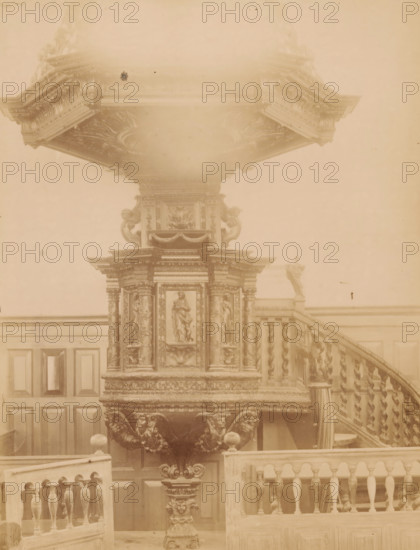 Pulpit in the Van Harenskerk in Sint-Annaparochie, photograph, annotation: 'St. Annaparochie, municipality of Het Bilt', annotation: 'Pulpit in the Dutch Reformed Church, in St. Anne parish, Friesland, Nov. 1898', Van Harenskerk, photographer: AJM Mulder (possibly), 1898, Sint Annaparochie, Attributed to, paper, pasteboard, albumen print, height 225 mm x width 171 mm, pulpit, ambo, chancel, A large ornate wooden pulpit occupying the center of the image with carved decorative panels and sculpted figures, a wide carved canopy above with layered moldings and intricate ceiling ornamentation, a spiral-carved staircase to the right with turned balusters and a small statue on the landing, paneled walls behind the pulpit, carved supports and ornamental lion-like figures beneath the pulpit, and a balustrade in the foreground.