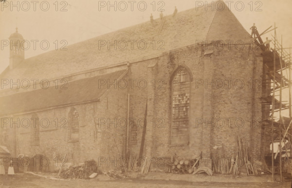 Broerekerk in Bolsward, during repair or restoration work, photograph, annotation: 'Bolsward (Fr), Broerenkerk, July 1903', annotation: 'Bolsward', Bolsward, photographer: AJM Mulder (possibly), 1903, Attributed to, paper, pasteboard, albumen print, height 151 mm x width 231 mm, church (exterior), A large brick building with a steep tiled roof fills the image, showing tall arched windows with broken or boarded panes, a small cupola near one end, and scaffolding against one corner where planks and beams are stacked; piles of wooden planks, beams, and stacked barrels lie along the base of the wall, and a ladder rests on the roof with several figures standing or sitting on the roof and near the scaffolding; the scene has a sepia tone and shows worn brickwork and scattered construction materials against a grassy foreground.