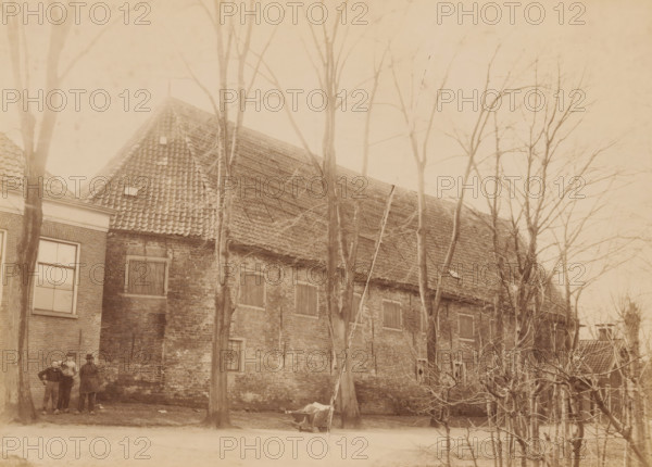 Begijnhofkapel te Bolsward, photograph, annotation: 'Bolsward', annotation: 'Bolsward, Begijnenhofkapel, naam 'de hok', april 1902', Bolsward, photographer: AJM Mulder (possibly), 1902, Attributed to, paper, pasteboard, albumen print, albumendruk, height 159 mm x width 221 mm, parts of church exterior and annexes: chapel, A sepia-toned photograph of a brick building with a steep tiled roof partially obscured by tall leafless trees, showing multiple shuttered windows and a larger window with panes on one side, bare branches in the foreground and a dirt road along the front, with a small group of three men standing near one tree and a wheelbarrow positioned near another tree.