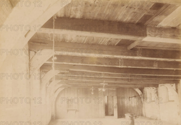 Interior of the Beguinage Chapel in Bolsward, photograph, annotation: 'Bolsward', annotation: 'Bolsward (Fr), Beguinage Chapel, Interior, April 1902', Bolsward, photographer: AJM Mulder (possibly), 1902, Attributed to, paper, pasteboard, albumen print, height 158 mm x width 229 mm, parts of church exterior and annexes: chapel, A high ceiling with exposed wooden beams and joists spans across the space, supported by large curved wooden brackets along the walls; a vertical rope hangs from one beam and several pendant light fixtures are suspended from the ceiling; wooden wall panels occupy the far wall with an open doorway and a small recessed window-like opening, and a freestanding cylindrical object with a rounded top sits near the foreground on the floor.
