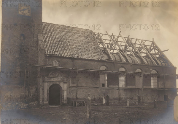 St. John the Baptist Church in Warns, during restoration work, photograph, annotation: 'Warns near Stavoren (Fr.), Dutch Reformed Church, Oct 1907', annotation: '..., Warns', Warns, photographer: AJM Mulder (possibly), 1907, Attributed to, paper, pasteboard, gelatin silver print, height 159 mm x width 225 mm, church (exterior), St. John the Baptist Church (Warns), A large brick building with a tall tower at one end occupies the scene, showing a steep tiled roof partially intact and partially removed to reveal a wooden roof framework and curved interior beams; the tower has a small arched window and a square clock face near its top. The facade features a central arched doorway with decorative stone surround above it and several tall arched windows along the wall, some with broken panes. Scaffolding and horizontal pipes run across the exterior, and wooden planks, construction debris, and upright posts are scattered on the ground in front of the building beneath a cloudy sky.