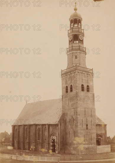Northwest side of the Great Church in Hindeloopen, photograph, annotation: 'Hindeloopen, Church of the Dutch Reformed congregation, NW side, March 1892', annotation: 'Hindeloopen', Hindeloopen, photographer: AJM Mulder (possibly), 1892, Attributed to, paper, pasteboard, albumen print, height 238 mm x width 169 mm, church (exterior), A sepia-toned photograph showing a tall brick church tower rising from a large church building with a steeply pitched roof; the tower has multiple tiers with arched openings, decorative railings, a domed cupola topped with a finial, and visible clock faces, while the lower tower shows arched windows and buttresses; the side of the church displays tall lancet windows and an entrance with an ornamented surround; a wooden fence runs along the foreground and small adjacent buildings are partially visible at the edges.