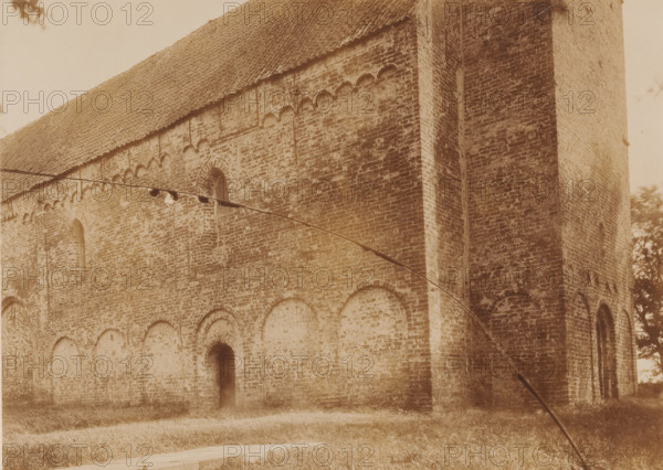 Sint-Martinuskerk in Westergeest, photograph, annotation: 'Kollumerland, Westergeest', annotation: 'Friesland, Kerk v, d NH gem. in Westergeest (Fr), Noordzijde, July 1895', Westergeest, photographer: AJM Mulder (possibly), 1895, Attributed to, paper, pasteboard, daylight collodion silver print, height 163 mm x width 224 mm, church (exterior), A sepia-toned photograph of a large brick building showing a sloped tiled roof and tall walls with decorative arched recesses and narrow arched openings, a vertical buttress or pilaster projecting from the right side, a dark narrow doorway set into the lower wall, grass in the foreground, a curved dark line crossing the image diagonally, and tree foliage visible at the image edges.