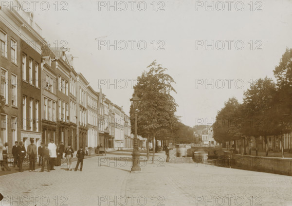 Street view in Leeuwarden, photograph, Leeuwarden, 1890 - 1920, photographer, paper, pasteboard, daylight collodion silver print, height 123 mm x width 174 mm, height 144 mm x width 198 mm, street (city(-scape) with figures, staffage), A wide cobblestone street runs alongside a canal with a stone bridge and row of trees, flanked on the left by a line of multi-story brick and stucco buildings with many tall windows and decorative facades, and on the right by dense tree foliage and more distant buildings; a tall ornate streetlamp stands near the canal edge, small boats sit on the water, and groups of people in period clothing stand and walk along the sidewalk near the buildings.