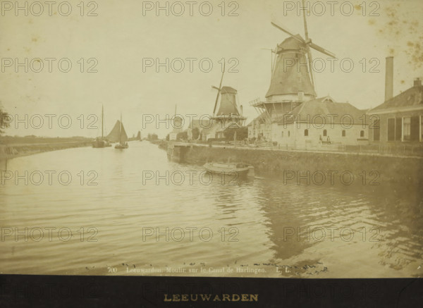Mills along the canal from Leeuwarden to Harlingen, photograph, inscription: '700 Leeuwarden. Moulin sur le Canal de Harlingen. LL', inscription: 'LEEUWARDEN', photographer: Léon & Lévy, Friesland, 1864 - 1900, paper, pasteboard, albumen print, albumendruk, height 120 mm x width 183 mm, height 139 mm x width 200 mm, canal, windmill in landscape, A wide canal scene with calm rippled water in the foreground and a shoreline with grass and a low embankment on the right, several boats moored and sailing on the water including a small rowboat near the right bank and larger sailboats further back, a row of traditional windmills of varying sizes along the right-hand side behind low buildings and fences, pitched roofs and chimneys on the buildings, a tall smokestack near the right edge, a few figures standing near the buildings by the water, and an open sky above.