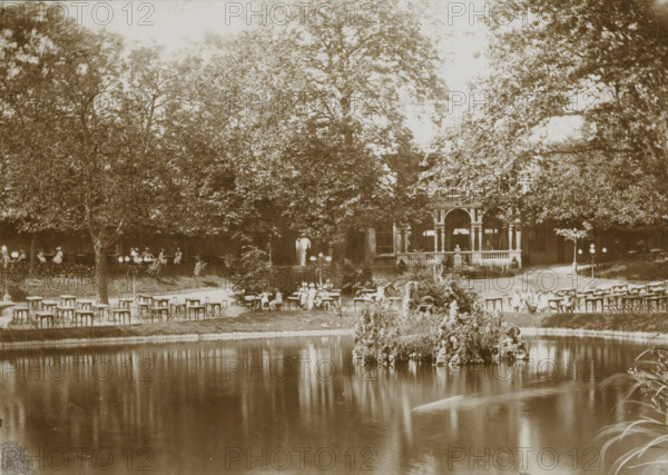 View of a park with a pond, presumably in Leeuwarden, photograph, Leeuwarden, 1890 - 1910, photographer, paper, pasteboard, daylight collodion silver print, height 124 mm x width 173 mm, height 144 mm x width 194 mm, park, A sepia-toned outdoor scene showing a pond in the foreground with a small rocky island covered in plants and flowers, its reflection visible in the water, and a grassy bank with scattered tables and chairs along the shore; beyond the seating area are large leafy trees partially obscuring a building with an ornate porch supported by columns, and people are seated at tables and standing near the structure, with lampposts and garden plantings interspersed among the trees.