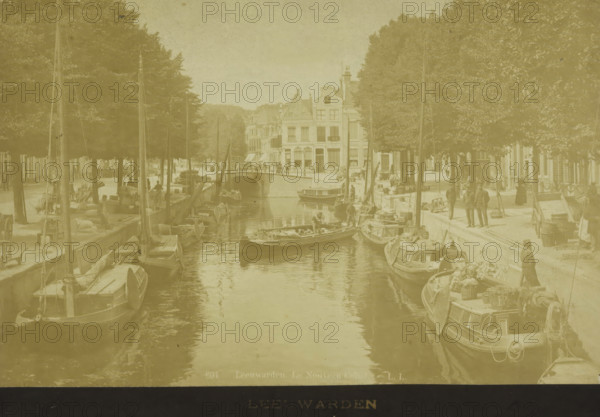Street view with canal in Leeuwarden, photograph, number: '... 50', retailer's mark: 'B. SMEDING, Boekhandel, LEEUWARDEN', inscription: 'LEEUWARDEN', inscription: '694 Leeuwarden. Le Nouveau Canal. LL', photographer: Léon & Lévy, Leeuwarden, 1864 - 1900, Léon & Lévy, photographer, paper, pasteboard, albumen print, height 121 mm x width 188 mm, height 140 mm x width 198 mm, canals, waters (in city), street (city(-scape) with figures, staffage), A canal scene with moored boats lining both sides of the water, tall masts rising above the vessels, and reflections visible on the water surface; a stone bridge spans the canal in the midground, beyond which a row of multi-story buildings with varied rooflines and windows faces the water; tree-lined quays run along both banks, with people standing and walking on the pathways beside the boats, and various small items and cargo placed near the vessels.