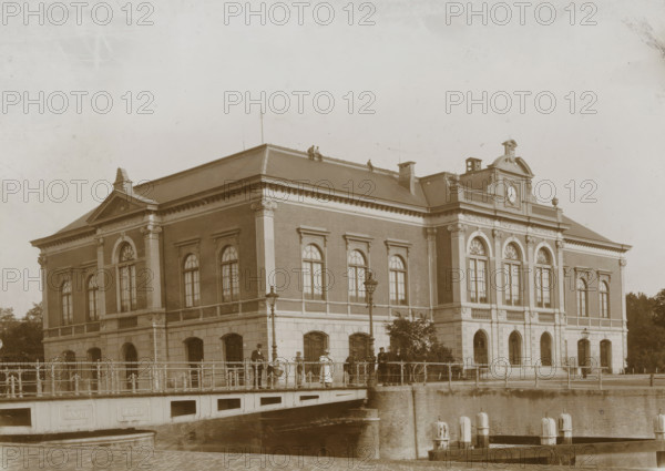 View of the former stock exchange building in Leeuwarden, photograph, Leeuwarden, 1890 - 1910, photographer, paper, pasteboard, daylight collodion silver print, height 122 mm x width 171 mm, height 144 mm x width 194 mm, Exchange, Bourse, A large stone and brick building with tall arched windows and decorative columns along its facade sits beside a canal, a sloped roof with small figures visible along the ridge, and a central pediment with a circular feature. A bridge with railings spans the water in front of the building, with lamp posts mounted along it and people standing and walking across, some wearing hats and long coats or dresses. The canal below the bridge has mooring posts and a stone retaining wall, and parts of the building's lower level show arched openings and a row of windows. The overall scene is in a monochrome sepia tone.