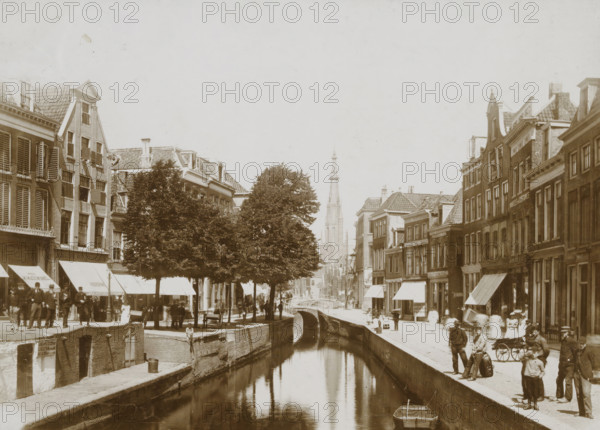 Street view with canal in Leeuwarden, photograph, Leeuwarden, 1890 - 1910, photographer, paper, pasteboard, daylight collodion silver print, height 122 mm x width 170 mm, height 144 mm x width 198 mm, canals, waters (in city), church (exterior), A canal runs down the center of the scene with calm reflective water and a small rowboat moored at the near edge, bordered by stone embankments and walkways. Lining both sides of the canal are narrow multi-story brick buildings with tiled roofs, shuttered windows, and awnings extending over the sidewalk. Several leafy trees stand near the left bank, partially obscuring some building facades. A stone pedestrian bridge arches over the canal further back, and beyond it a tall ornate church tower rises against a pale sky. Numerous people occupy the sidewalks and embankments wearing period clothing, with some standing in groups, some seated, and a few children visible near the foreground. Benches, carts, and street lamps are present along the walkwa...