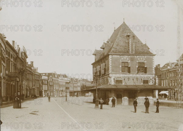 Street view with the Waag in Leeuwarden, photograph, photographer: anonymous, Leeuwarden, 1890 - 1910, paper, pasteboard, daylight collodion silver print, height 126 mm x width 174 mm, height 144 mm x width 199 mm, street (city(-scape) with figures, staffage), weigh-house, A wide cobblestone street lined with multi-story brick buildings leads to a large steep-roofed brick building with dormer windows and a covered arcade at its base; people stand and walk in the open area in front of the building, including men in dark suits and hats and a woman in a long dark dress with a hat, while lamp posts and a few small street-level awnings are visible along the street and façades of adjacent buildings.
