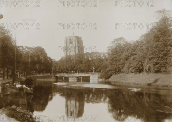 View of the Oldehove with canal in Leeuwarden, photograph, Leeuwarden, 1890 - 1910, photographer, paper, pasteboard, daylight collodion silver print, height 125 mm x width 173 mm, height 144 mm x width 194 mm, canals, waters (in city), parts of church exterior and annexes: tower, A sepia-toned riverside scene showing a tall stone tower rising above a row of trees, a low bridge crossing the river in front of the trees, reflections of the tower and trees visible on the water surface, boats moored along the near bank with some rigging and equipment, dense foliage on both sides of the river, a lamp post near the bridge, and a calm sky overhead.
