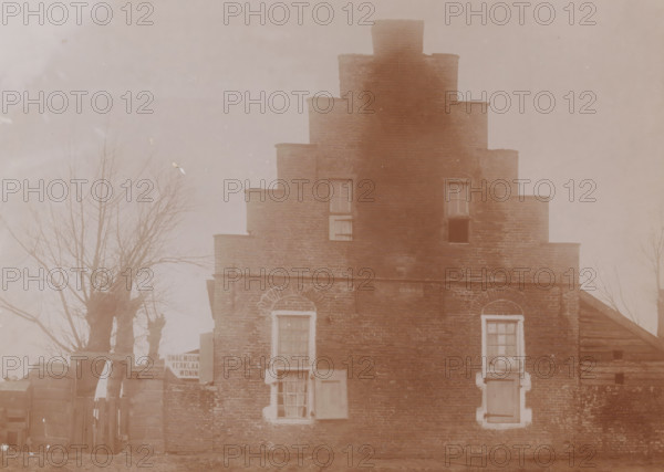 Aytta State (godshouse or church) in Swichum, photograph, annotation: 'Aytta Godshuis Swichem, Friesland, Dec. 1909, Leeuwarderadeel', Swichum, photographer: AJM Mulder (possibly), 1909, Attributed to, paper, pasteboard, daylight collodion silver print, height 151 mm x width 209 mm, church (exterior), A sepia-toned photograph of a stepped brick building with a central raised section and tiered sides, showing multiple windows with shutters and panes in varying states, two arched brick window surrounds on the lower level, and a doorway partially visible. Bare tree branches flank the building on both sides, and a wooden fence and gate are visible at the left base of the building. The brickwork shows weathering and variations in tone, and the overall scene has a faded, aged appearance.