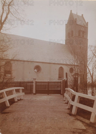 St. John the Baptist Church in Huizum, photograph, annotation: 'Dutch Reformed Church with tower in Huizum (Fr) Jan 1911, Leeuwarderadeel', Huizum, photographer: AJM Mulder (possibly), 1911, Attributed to, paper, pasteboard, daylight collodion silver print, height 216 mm x width 151 mm, church (exterior), A wooden bridge with painted railings leads to a gate with vertical slats and posts, beyond which stands a building with a long sloped roof and smooth walls. The building facade shows rounded and arched windows, one circular window with visible radial mullions and a taller arched window nearby. To the right a tall brick tower with arched openings and a steep roof rises above the building. Bare tree branches frame the scene on both sides and a pale sky fills the upper background.