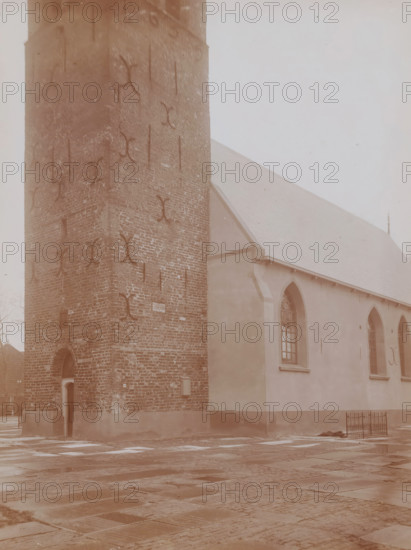 St. John the Baptist Church in Huizum, photograph, annotation: 'Detail of tower and Church of the Dutch Reformed Congregation in Huizum (Fr) Jan 1911, Leeuwarderadeel', Huizum, photographer: AJM Mulder (possibly), 1911, Attributed to, paper, pasteboard, daylight collodion silver print, height 218 mm x width 163 mm, church (exterior), A tall brick tower with visible metal anchors and an arched doorway sits adjacent to a plastered building with arched windows and a sloped roof, the ground in front paved with large rectangular stones and wet patches, and a small metal fence near the building's corner.