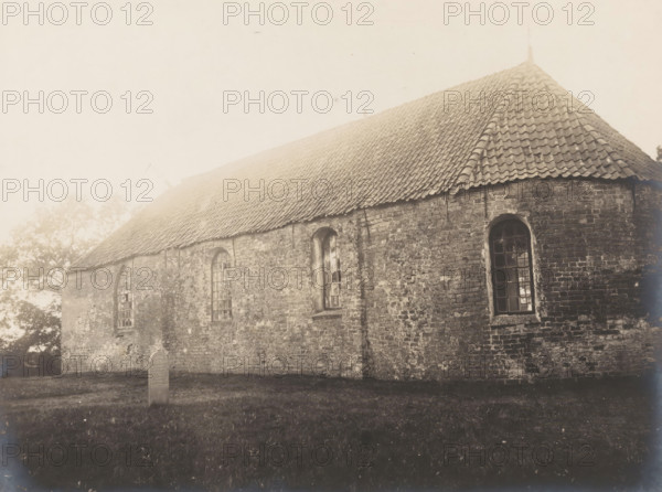 View of the Reformed Church in Siegerswoude, demolished in 1909, photograph, annotation: 'Siggerswoude, Interior view, looking east, Oct 1908, Opsterland', Siegerswoude, photographer: AJM Mulder (possibly), 1908, Attributed to, paper, pasteboard, gelatin silver print, developing gelatin silver print, height 162 mm x width 216 mm, church (exterior), A long brick building with a sloped tiled roof fills the frame, showing a series of tall arched windows set into the weathered brick wall, one of which has visible windowpanes and reflections, while the roof displays rows of overlapping tiles and a pointed finial at the far end; a patch of grass lies in the foreground and a solitary upright stone marker stands near the building, with trees faintly visible behind the left side of the structure.