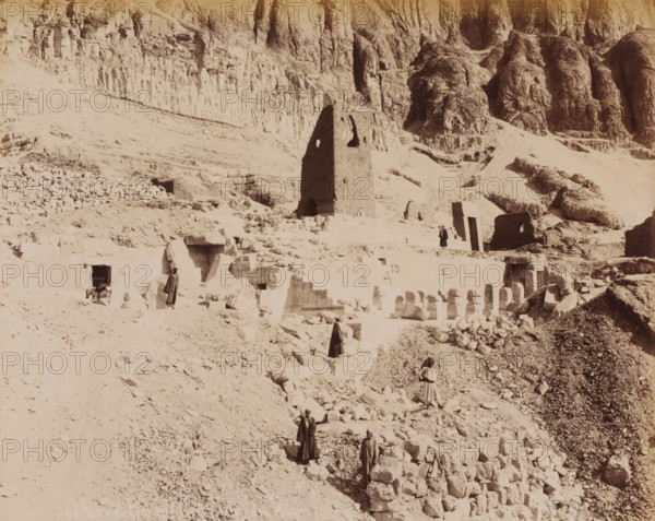 Ruins of Dar-el-Bahar, Safi, Morocco, album page, photograph, Dar-el-Bahar is a Portuguese fortress (from the time of Portuguese rule from 1488 to 1541) in the city of Safi on the west coast of Morocco., inscription: 'Der-el-Bahar (Vue des Ruins)', number: '744', c. 1880 - c. 1900, photographer, pasteboard, paper, albumen print, albumendruk, height 270 mm x width 214 mm, height 266 mm x width 357 mm, Landscape format, gelding, North Africa, A sepia-toned desert scene showing a rocky hillside with carved stone structures and ruins, including a tall, weathered stone tower with openings, terraces of cut blocks, scattered rubble and boulders, steep eroded cliffs in the background, several people wearing long robes standing or walking among the stones, and a donkey near a dark entrance set into the rock.