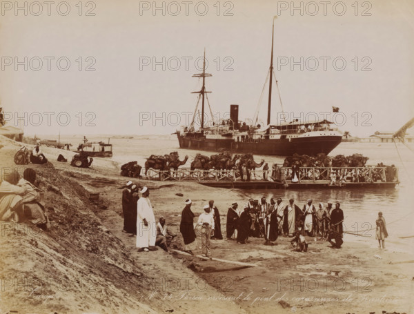 Station in the Suez Canal, Kantara, Egypt, photograph, View of a mooring place and of the Suez Canal with a ship on the right and camels on the gangplank., number: '1710', inscription: 'Zangaki', inscription: 'No. 74 Persia croisant le pont des caravannes de Kantara', photographer: Zangaki, Cairo (city), c. 1880 - c. 1900, Ottoman Empire, North Africa, paper, albumen print, height 219 mm x width 285 mm, Suez Canal, A large steamship with tall masts and a smokestack is moored near a wooden pier, with a flag flying from its stern; a number of camels loaded with packs stand on and near the pier while groups of people in robes and head coverings gather on the shore and on the pier, some standing and some sitting; a smaller boat floats beside the shore and wooden structures and low buildings are visible in the background along the waterline; the foreground shows a sloping sandy embankment with seated and standing figures and scattered items on the ground.