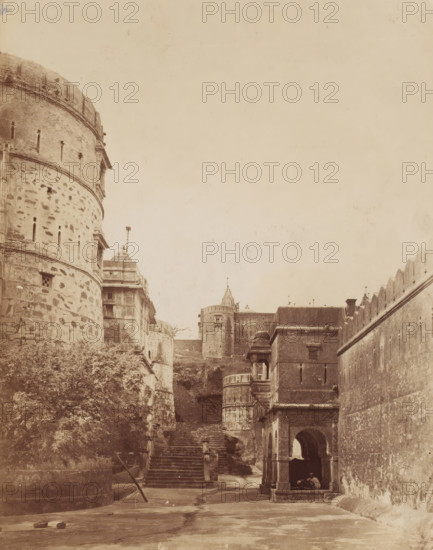 Temple complex at Palitana, Gujarat, India, photograph, Castle complex ?, India ?., Palitana, c. 1870 - c. 1900, photographer, paper, albumen print, height 232 mm x width 183 mm, A sepia-toned view of a stone fortress complex with rounded towers and high walls; a large cylindrical tower with small vertical slits and textured stonework occupies the left foreground, while a series of steps lead uphill between buildings toward additional towers and battlements in the midground; on the right a tall wall with crenellations runs diagonally, and nearer to the viewer a square stone pavilion with arched openings and a domed roof sits beside the steps; vegetation grows at the base of the left tower and two small objects lie on the ground in the open courtyard area in the foreground.