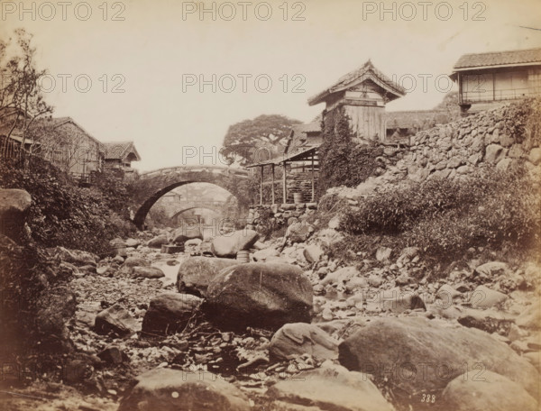 The Amidabashi Bridge and behind it the Koraibashi Bridge over the Nakashimagawa River, Nagasaki, Japan, photograph, The photo was taken downstream. Nearby was the house of Ueno Hikoma. See Nagasaki University Library Collection, where the same photo (colored)., number: '388', inscription: 'illegible', c. 1870 - c. 1900, photographer, paper, albumen print, height 195 mm x width 258 mm, Kyushu, A sepia-toned scene shows a rocky streambed extending toward the background with smooth boulders and smaller stones scattered along the water and banks, wooden tubs and buckets resting among the rocks, a stone arched bridge crossing the stream with a second distant arch visible beyond it, timber and stone buildings with tiled roofs lining the banks including a tall wooden structure partially covered in climbing vegetation, a low wooden shelter with posts near the water, a stone retaining wall supporting a slope on one side, patches of grass and shrubs on the banks, and a large tree with a broad c...