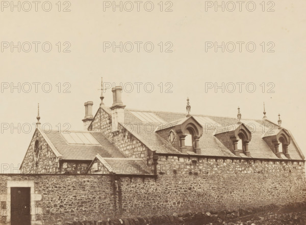 View of country house in England, photograph, England, c. 1875 - c. 1885, photographer, pasteboard, paper, albumen print, albumendruk, height 196 mm x width 262 mm, height 402 mm x width 582 mm, farm or solitary house in landscape, A sepia-toned view of a stone building with a tiled roof and multiple gables, featuring decorative roof finials and chimney stacks, dormer windows with arched tops and columns, rectangular skylights set into the roof, and a high rough stone wall in the foreground with a dark wooden door set into a lighter stone frame.