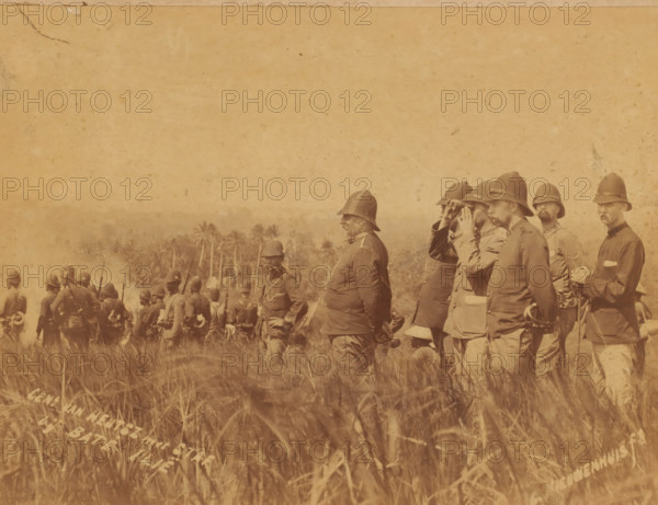 General van Heutsz with staff in Aceh during the attack on the benteng complex Batoe Iliq in the coastal state of Samalanga, photograph, General van Heutsz with staff in Aceh., inscription: 'Gen. van Heutsz with staff, at Baté Illië, C. Nieuwenhuis ft.', photographer: Christiaan Benjamin Nieuwenhuis, Aceh, 1901-03-12, Sumatra, pasteboard, paper, albumen print, albumendruk, height 218 mm x width 284 mm, height 326 mm x width 440 mm, Person, Johannes Benedictus van Heutsz, commander-in-chief, general, marshal, binoculars, spy-glass, A sepia-toned photograph showing a group of men in military uniforms and pith helmets standing and facing toward the left, some holding rifles and one using binoculars, with tall grasses in the foreground and a line of palm trees visible in the middle background.
