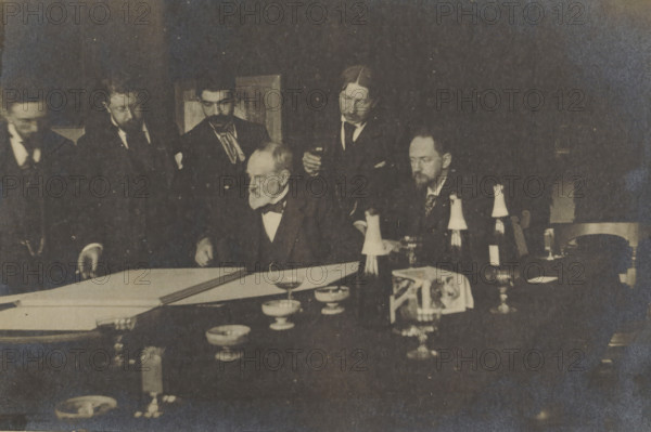 George Hendrik Breitner and others, looking at a portfolio, possibly during an art viewing at Arti et Amicitiae, photograph, Amsterdam, 1900 - 1923, photographer, paper, pasteboard, baryta paper, gelatin silver print, developing gelatin silver print, height 153 mm x width 227 mm, Person, George Hendrik Breitner, historical persons, painter in non-work situation (circle, club of artists), Arti et Amicitiae, A dim, sepia-toned scene of men gathered around a table with documents and tableware; several men in dark suits and bow ties stand and sit closely together, one older man with a white beard seated near the center leaning over a large sheet on the table while others look on, some holding small glasses; the table foreground shows bowls, goblets, bottles with paper coverings, a folded cloth with an image, and scattered small dishes and glassware, with a chair partially visible at the right and a framed object on the wall behind the group.