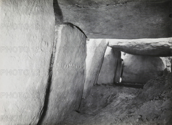 Hunebed van Kerguntuil, Trégastel, photograph, inscription: 'IX, Ban v Heuvel, Allée Couverte de Kerguntuil, Trégastel C. du N., sculpture on two supporting stones', photographer: attributed to Jan Lanting, supervision: Albert Egges van Giffen, 1939-05-27 - 1939-06-04, Attributed to, paper, baryta paper, gelatin silver print, developing gelatin silver print, height 123 mm x width 172 mm, megalithic chambered tomb, dolmen, A close-up interior view of a narrow stone passage showing rough, weathered vertical slabs forming the sides and flat horizontal slabs forming the ceiling, with visible stone textures, joints, and chipped edges; the ground is uneven and covered in compacted dirt and small debris; light enters from an opening at the far end, illuminating the irregular surfaces and casting soft shadows along the stones.