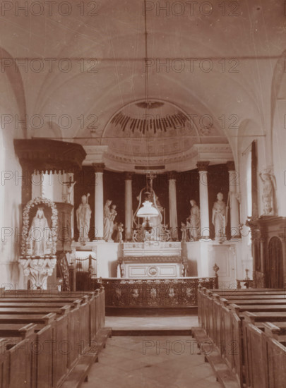 Interior of the St. Lambertus Church in Huisseling, photograph, annotation: 'Municipality of Ravenstein, Roman Catholic church in Huiselinge (NB) near Ravenstein March 1911', Huisseling, 1911, photographer, Attributed to, paper, pasteboard, daylight collodion silver print, height 218 mm x width 161 mm, interior of church, A sepia-toned interior view of a church nave with wooden pews leading toward an ornate altar area, flanked by tall columns and statues on raised pedestals; a domed ceiling with decorative molding is visible above the altar, and a hanging lamp or chandelier is suspended in front of the altar; carved woodwork, decorative panels, and sculptural figures, including a draped statue in a floral arch on one side, are arranged symmetrically around the central altar; railings and a patterned altar frontal occupy the foreground, and high arched walls and windows frame the scene.