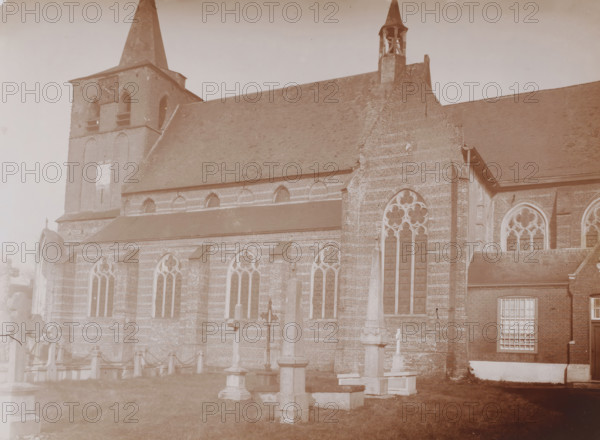 South side of the St. Lambertus Church in Rosmalen, photograph, annotation: 'Rosmalen, March 1910', Rosmalen, 1910, photographer, Attributed to, paper, pasteboard, daylight collodion silver print, height 159 mm x width 221 mm, churchyard, cemetery, church (exterior), A large brick church with tall sloped roofs and two steeples dominates the scene, showing pointed arched windows with tracery and stained glass along its side, buttresses and decorative brickwork, and a tower with openings and a clock face. In front of the churchyard are several stone crosses and tomb markers on plinths connected by short posts and chains, set on a grassy area. The image has a sepia tone and shows part of an adjacent brick building with a multi-paned window, along with worn masonry and weathered surfaces on the church walls.