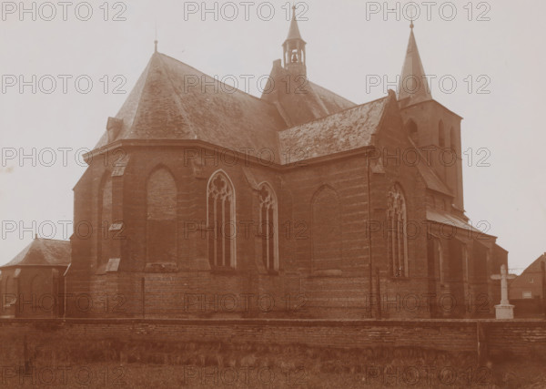 Northeast side of the St. Lambertus Church in Rosmalen, photograph, annotation: 'Rosmalen, March 1910', Rosmalen, 1910, photographer, Attributed to, paper, pasteboard, daylight collodion silver print, height 158 mm x width 221 mm, church (exterior), A large brick church building fills the image, showing steep pitched roofs covered in shingles, pointed spires, a small open bell tower, and tall narrow arched windows with tracery. The exterior walls display buttresses and recessed arched panels, and a low brick wall runs along the foreground with grass and vegetation in front of it. The overall tone of the photograph is sepia, and the sky appears pale and empty above the building.