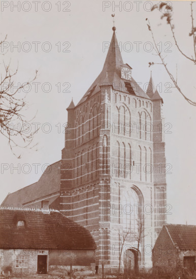 Tower of the Reformed Church in Sprang-Capelle, photograph, annotation: 'Tower of the NH church in Sprang (NB) after the restoration 1911', annotation: 'J. Kuiper (practice)', Sprang-Capelle, in or after 1911 - c. 1920, photographer, Attributed to, paper, pasteboard, daylight collodion silver print, height 220 mm x width 155 mm, parts of church exterior and annexes: tower, A tall brick church tower with arched window openings and vertical stone banding rises behind smaller thatched and tiled roof buildings, the tower topped by a steep spire with a clock face and finial, bare tree branches frame the scene on both sides, and a wooden arched doorway and narrow windows are visible at the tower base.