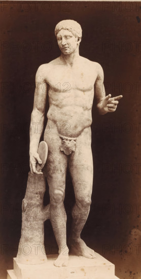 Sculpture of a discus thrower, Museo Vaticano, photograph, photographer: Libreria Spithöver, c. 1850 - c. 1870, pasteboard, paper, albumen print, height 383 mm x width 200 mm, piece of sculpture, reproduction of a piece of sculpture, Vatican City, Vatican Museums, A life-size marble statue of a nude male figure standing on a rectangular plinth, shown against a dark background, with a relaxed contrapposto pose; the figure's left arm is bent and the hand extended with two fingers pointing, while the right arm hangs down holding a rounded object that rests against a vertical support resembling a tree trunk; the head is turned slightly to the left with short curly hair and a calm facial expression, and a small decorative covering is placed over the groin; the surface of the sculpture shows weathered texture and subtle discoloration, and the plinth bears a small rectangular label centered on its front edge.