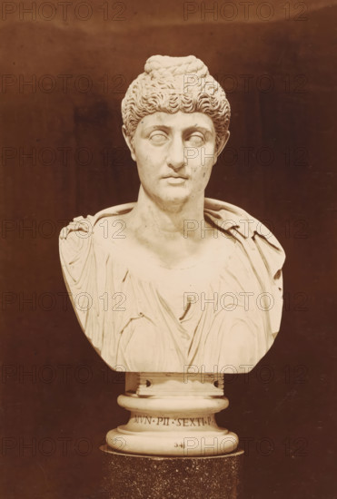 Sculpture (bust) of Faustina, Museo Vaticano, photograph, photographer: Libreria Spithöver, c. 1850 - c. 1870, pasteboard, paper, albumen print, height 334 mm x width 228 mm, piece of sculpture, reproduction of a piece of sculpture, Vatican City, Vatican Museums, A white marble bust of a person with short curly hair gathered into a braided bun on top, featuring a neutral expression with detailed facial features and smooth polished surfaces, draped clothing with carved folds across the shoulders and chest, and a round pedestal base with carved decorative rings and an inscription on its front, set against a plain dark brown background.